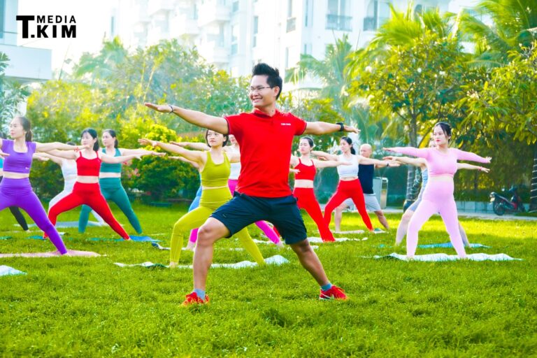 Đặng Kim Ba mặc áo đỏ dẫn một lớp Yoga ngoài trời đông người trên bãi cỏ, cả lớp đang ở tư thế chiến binh hai
