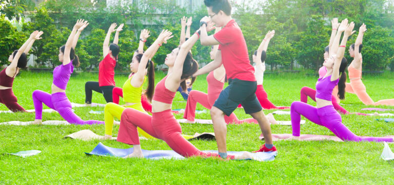Đặng Kim Ba đứng giữa hướng dẫn một lớp Yoga ngoài trời, học viên tập tư thế chiến binh trên thảm giữa bãi cỏ