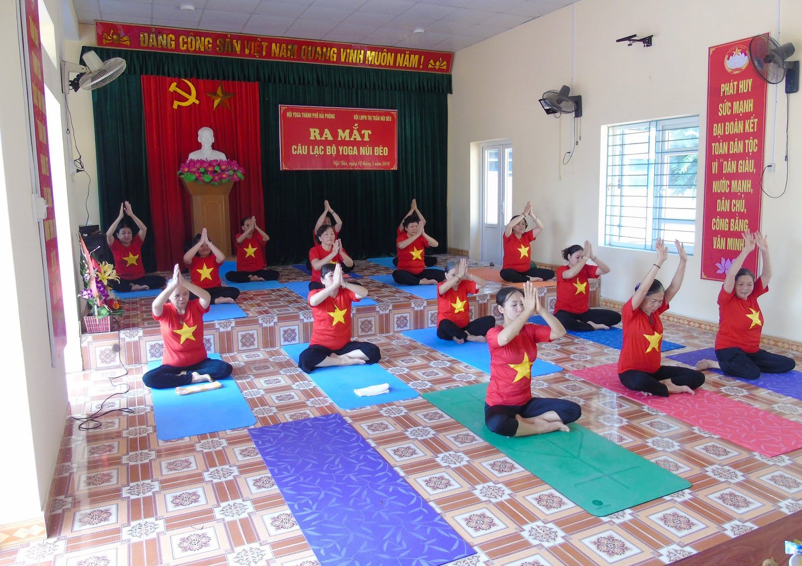 Lớp Yoga của một câu lạc bộ thị trấn tập trong hội trường nhà văn hoá: sàn gạch hoa, quạt treo tường, mỗi người một tấm thảm màu, phía trên sân khấu là băng rôn ra mắt câu lạc bộ