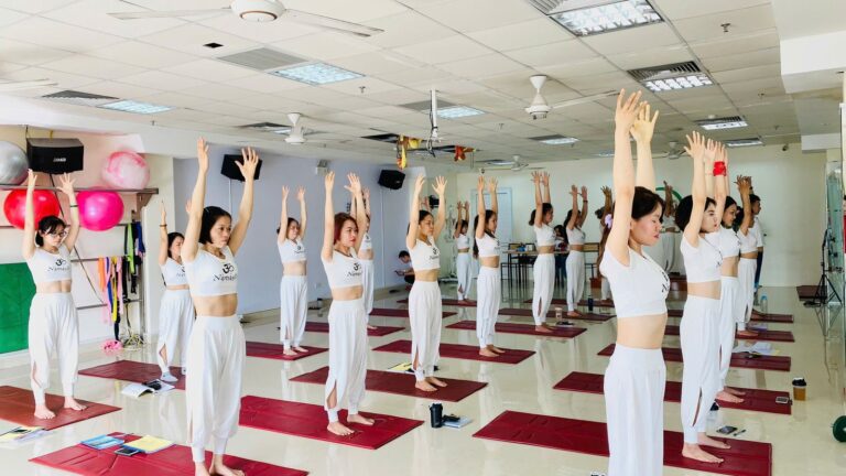 Lớp Yoga trong một phòng tập bình dị, học viên mặc đồng phục trắng đứng trên thảm đỏ, cùng vươn hai tay lên cao